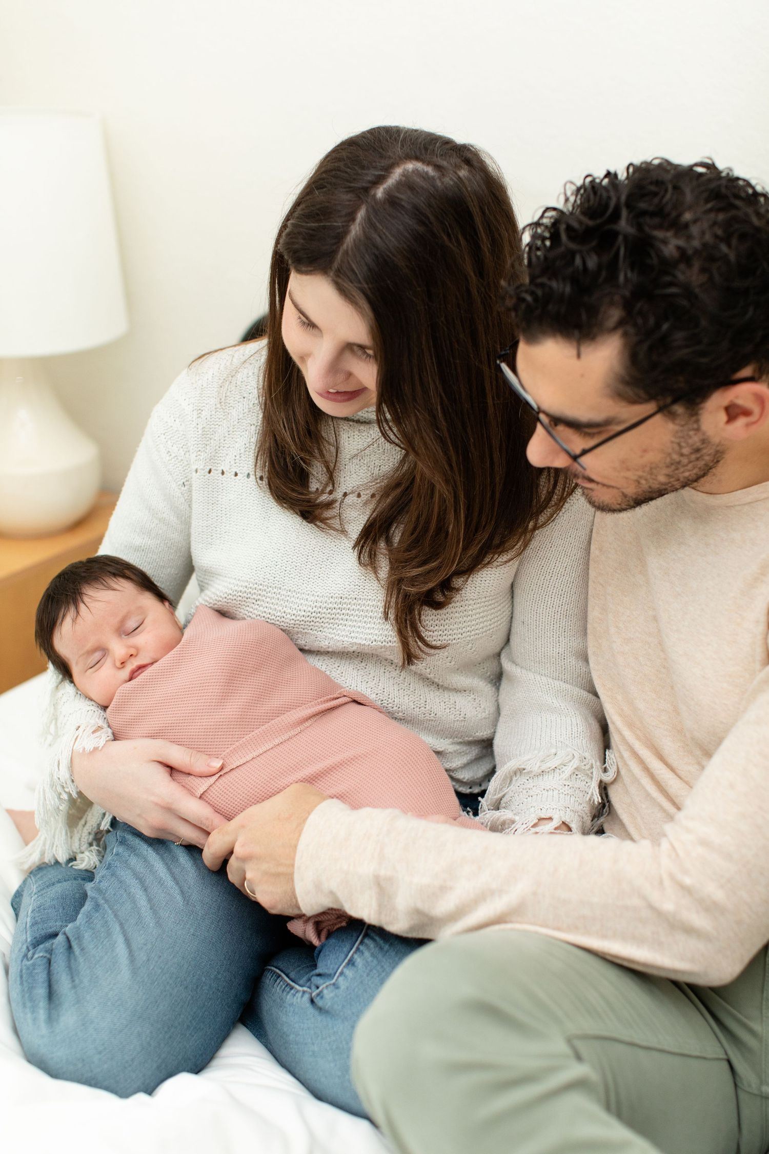 Parents gaze lovingly at their sleeping newborn baby wrapped in pink on white bedding.