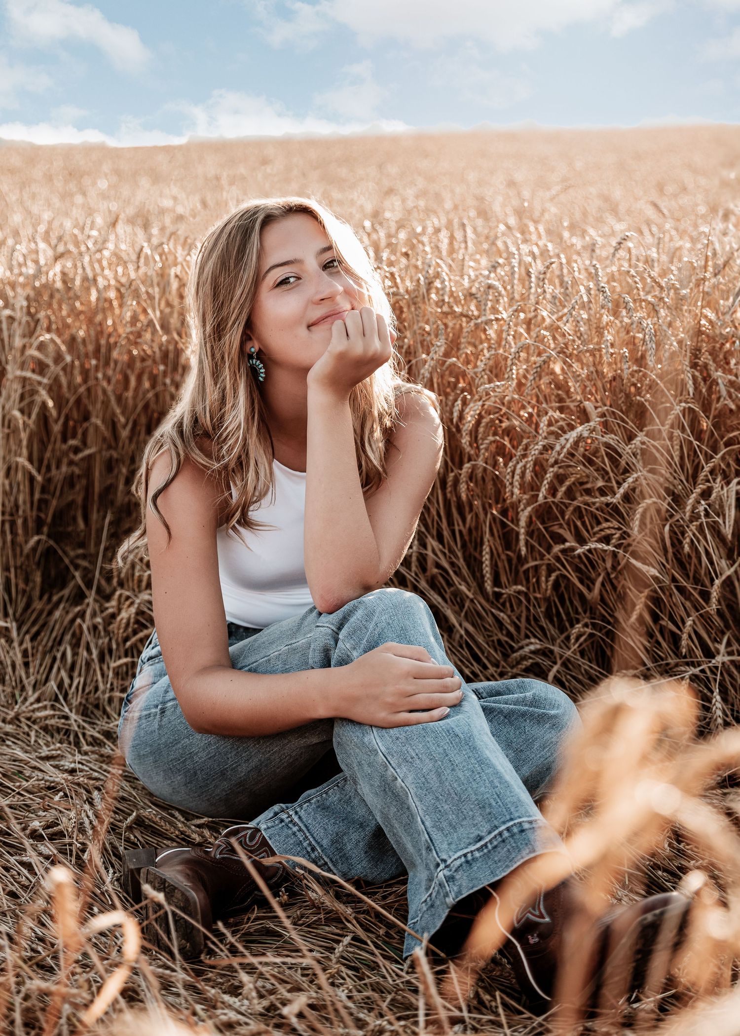 Woman in white top and jeans sits thoughtfully in golden wheat field at sunset.