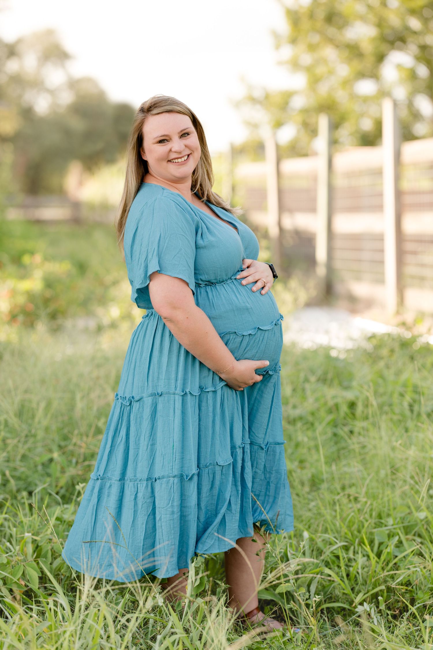Expectant mother in blue tiered dress poses in outdoor garden setting.