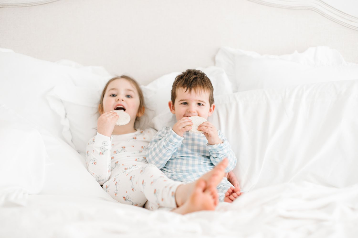 Children in pajamas playing and laughing together on white bedding.