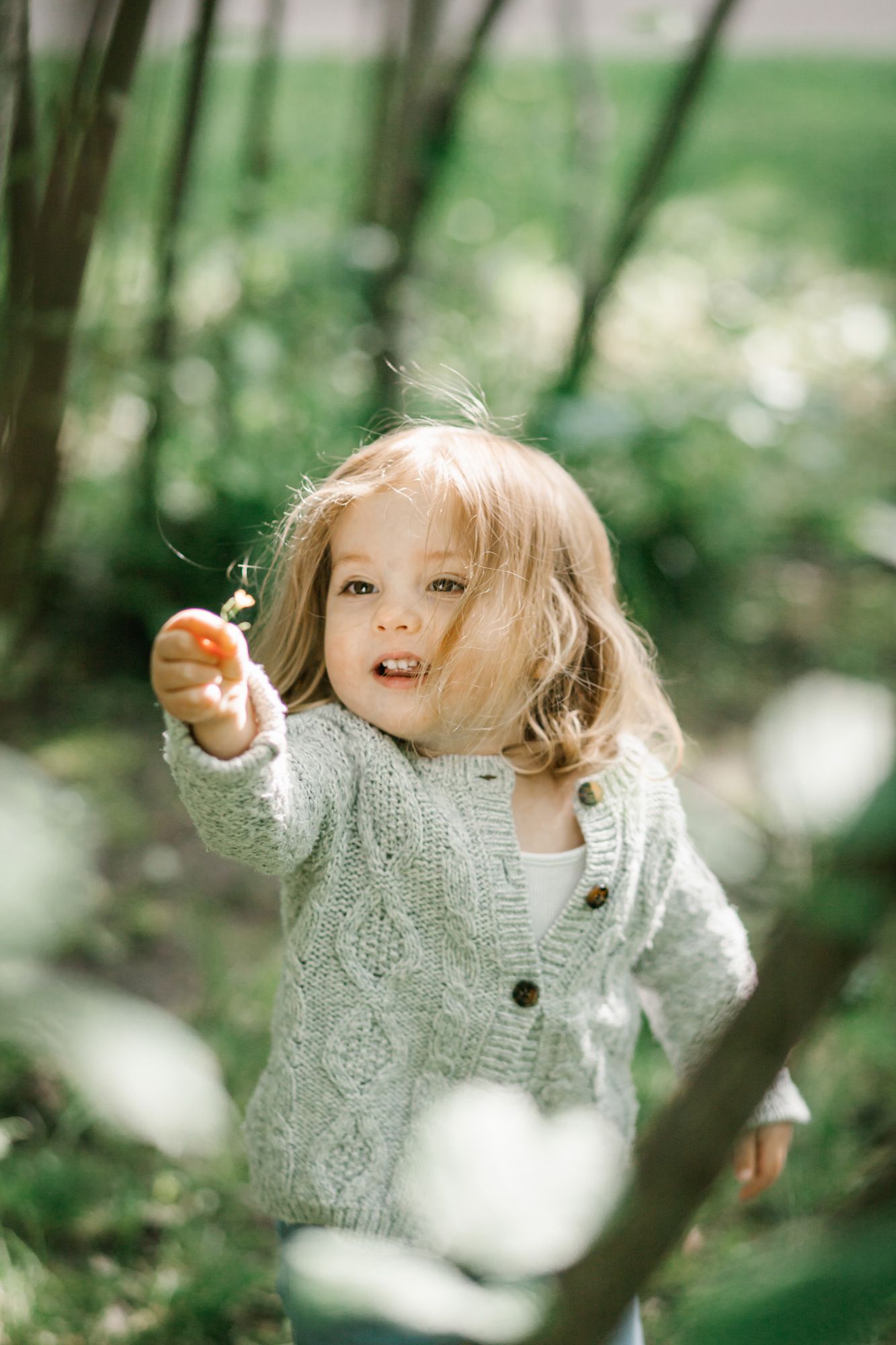 A small child in a knit sweater points while playing outdoors in a dreamy forest setting.