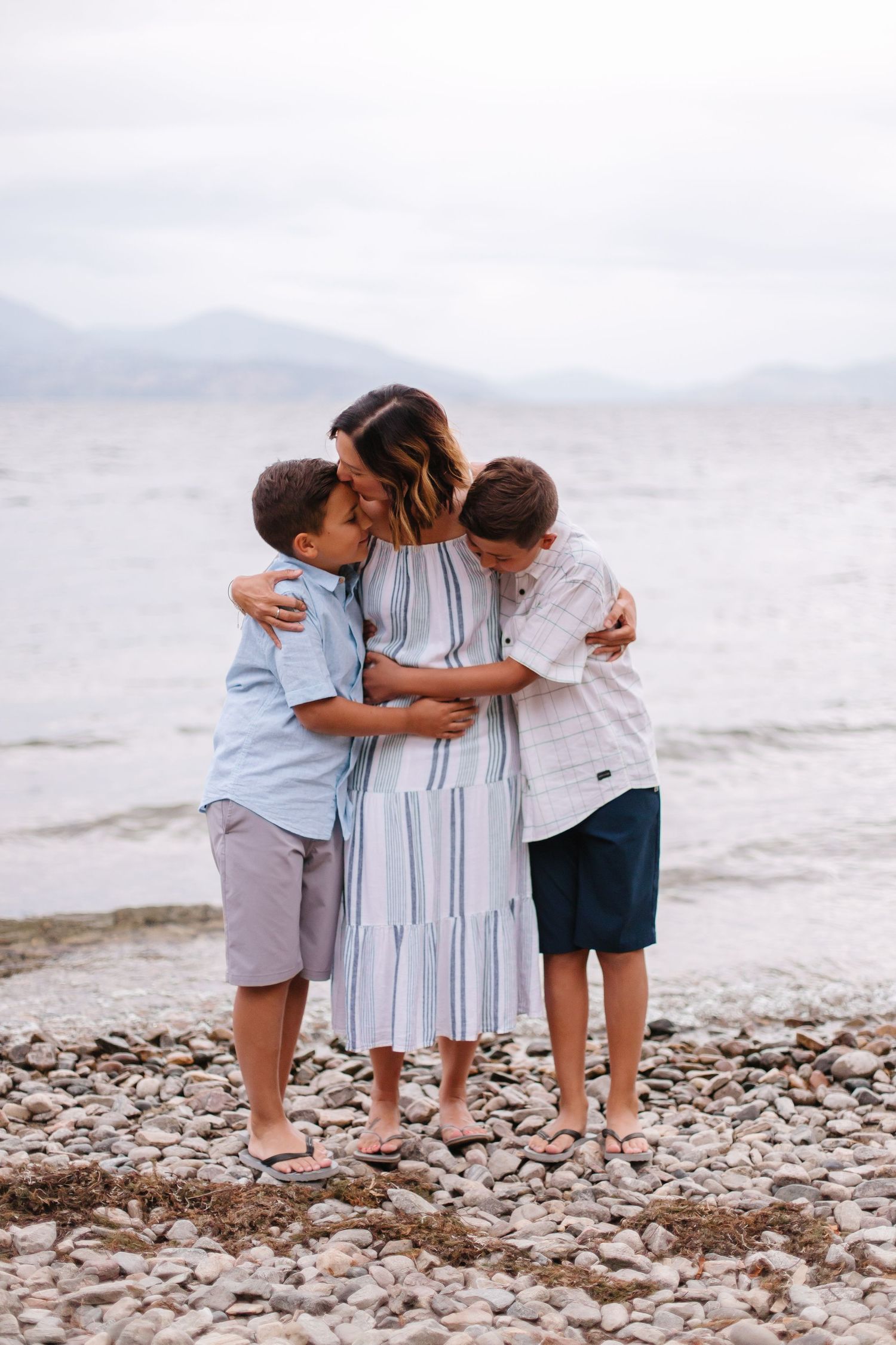 Three people embrace on a pebble beach with waves in the background.
