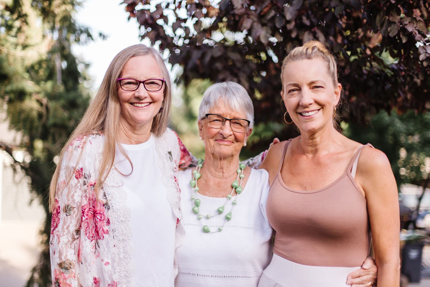 Three generations of women smile together outdoors under trees.