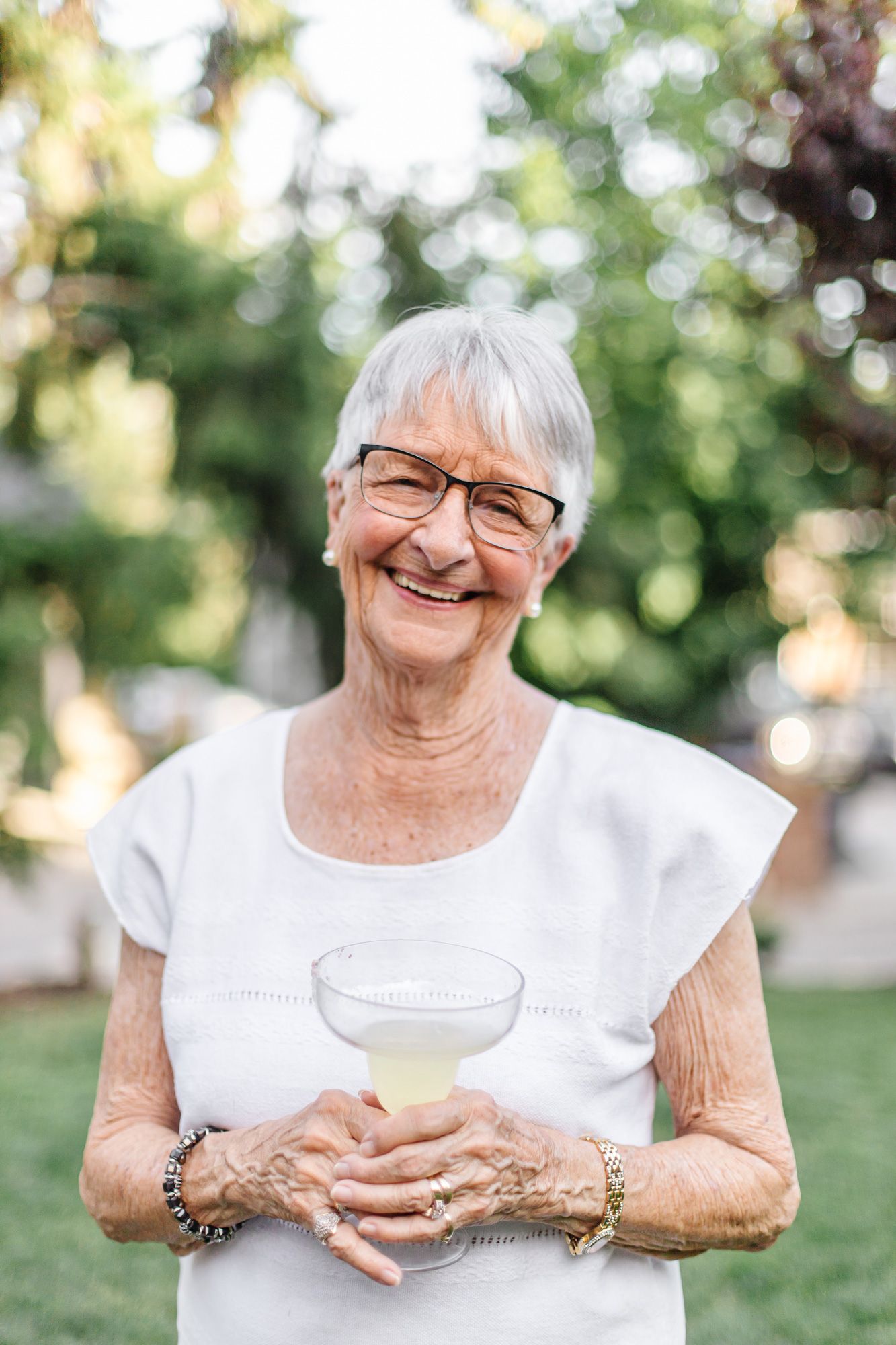 Person in white top holding cocktail glass smiles outdoors with greenery in background.