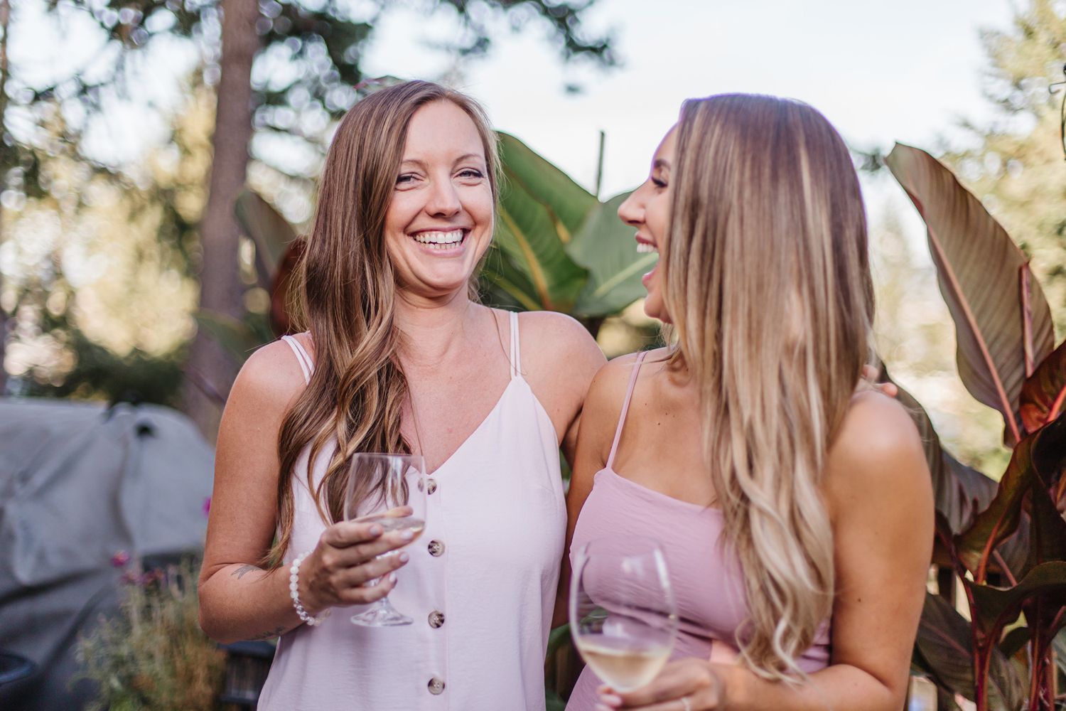Two friends laughing together while holding wine glasses at outdoor gathering.