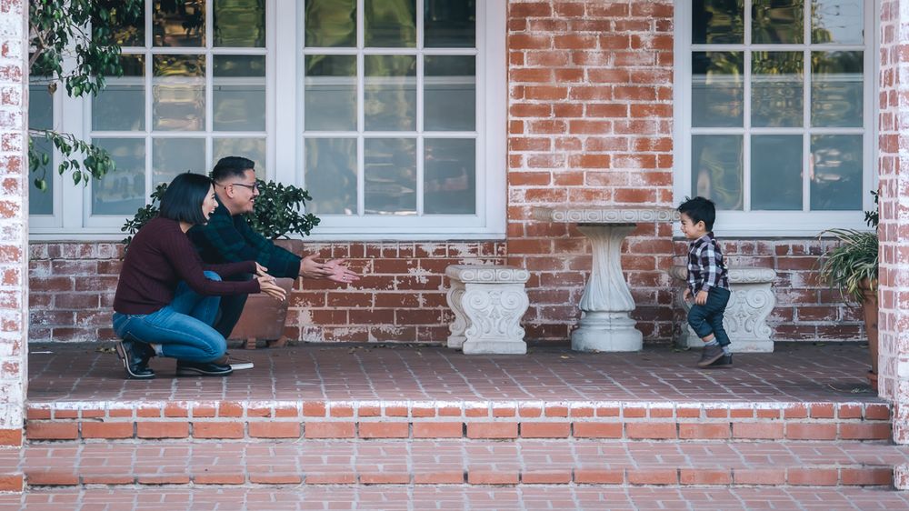 A man crouches down to interact with a young toddler on a brick home's front porch.