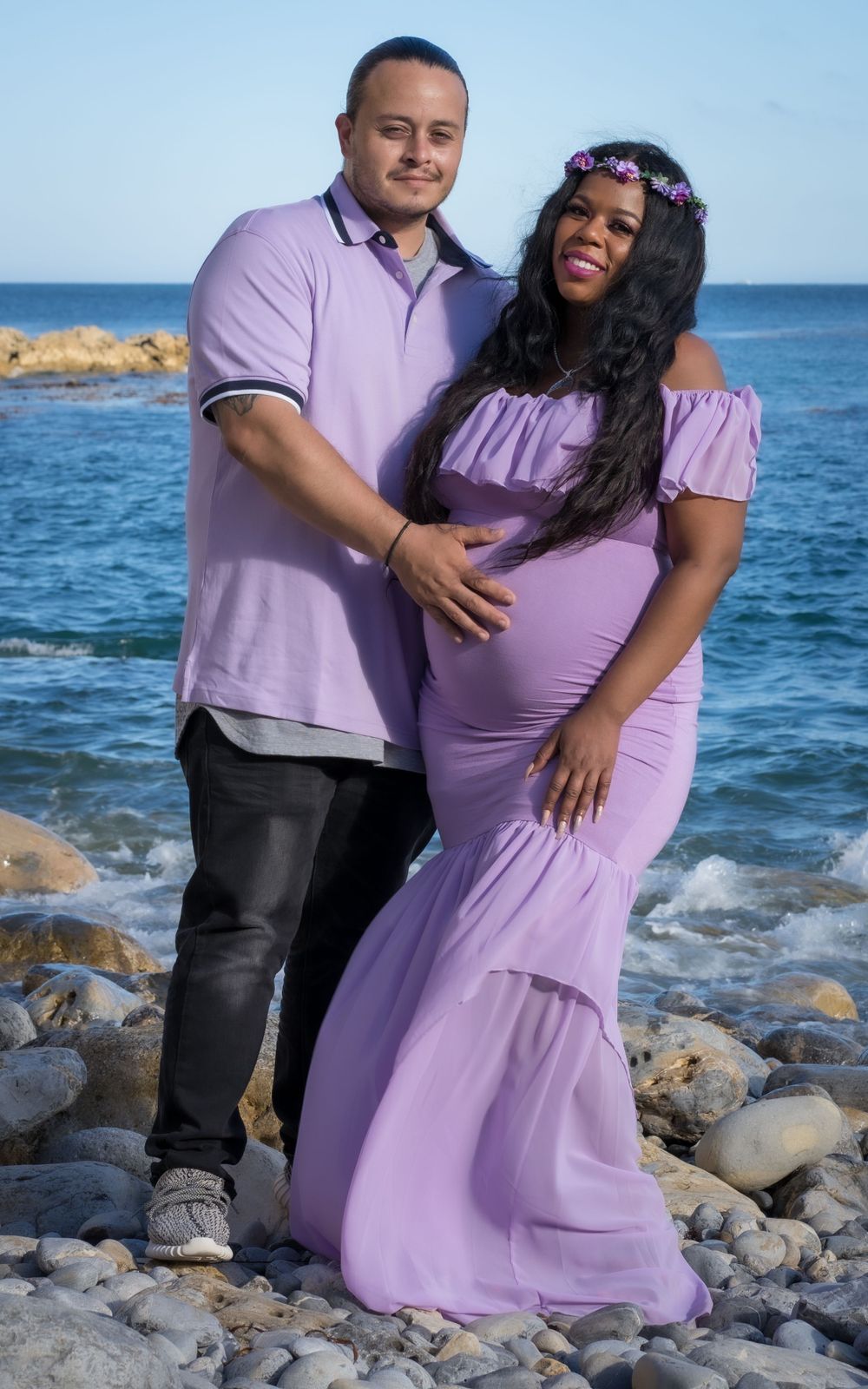 Expecting couple in matching lavender outfits pose together on a rocky ocean shoreline.