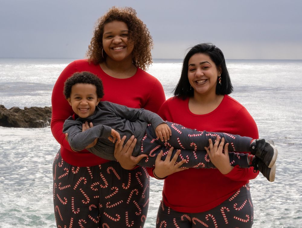 Two women in matching red and candy cane print holiday pajamas hold a baby boy near the ocean.