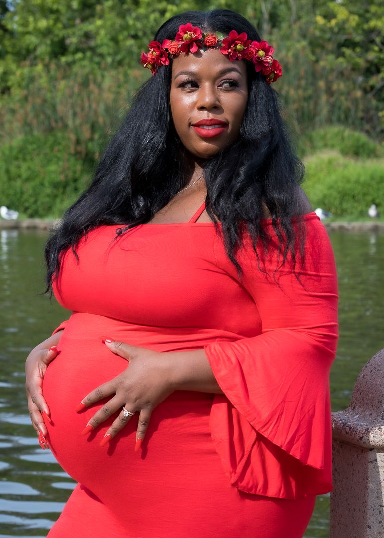 Pregnant Black woman in red off-shoulder dress with floral crown poses near a pond.