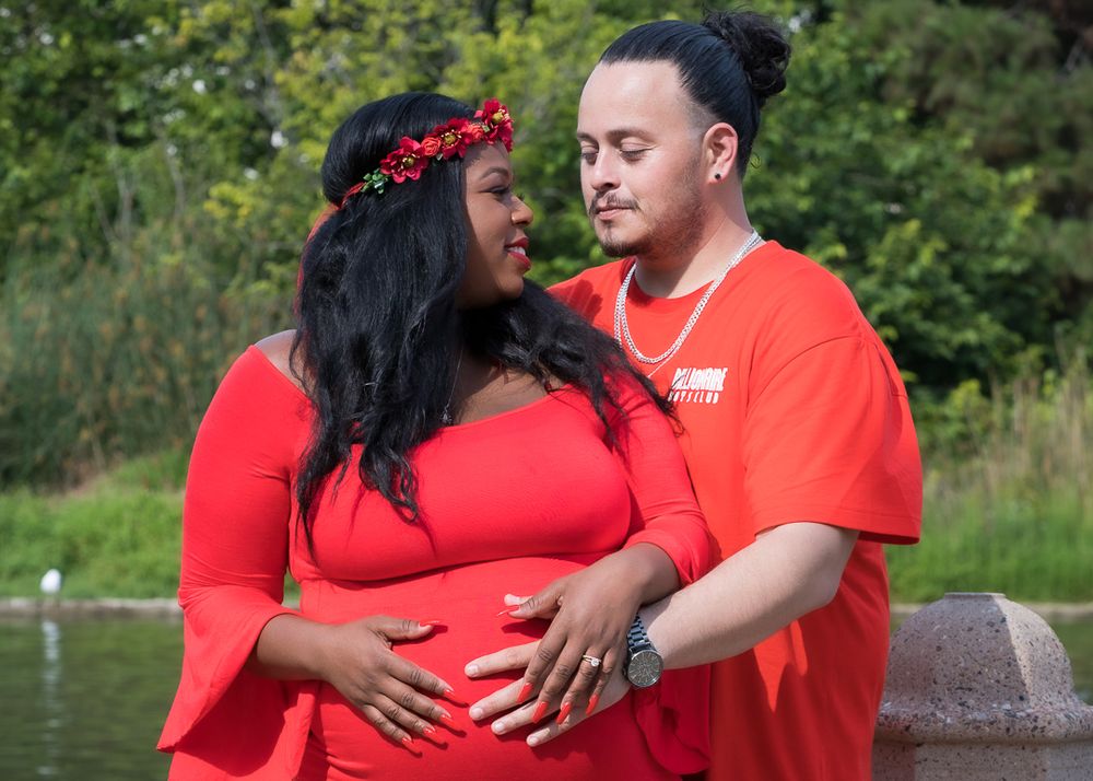 Expecting couple in matching red outfits tenderly embrace near a peaceful pond outdoors.