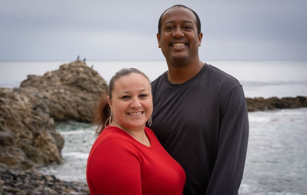 Smiling couple in red and black tops embracing near a rocky ocean shore.