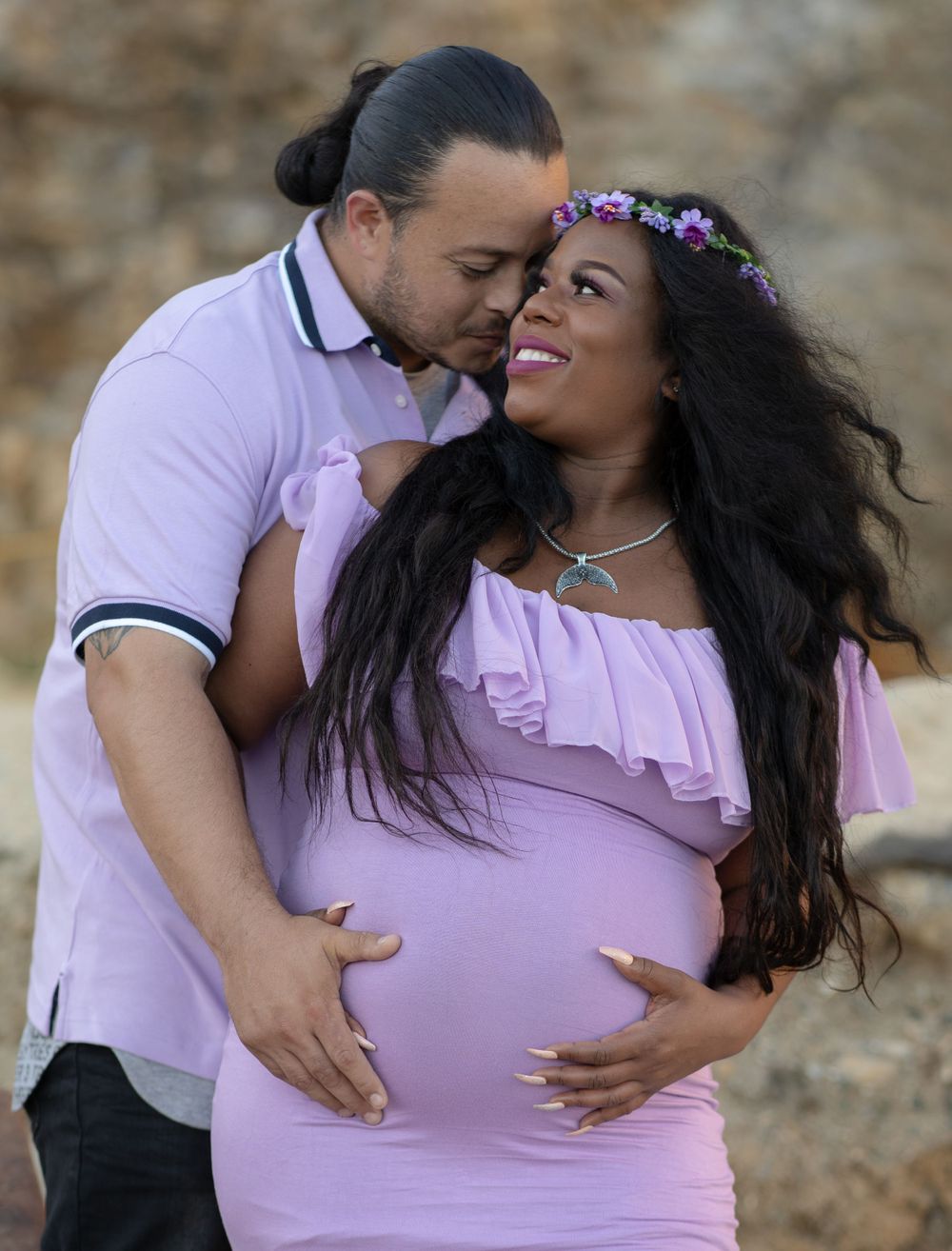 Loving couple embraces on rocky beach, man kisses pregnant woman wearing purple floral crown.