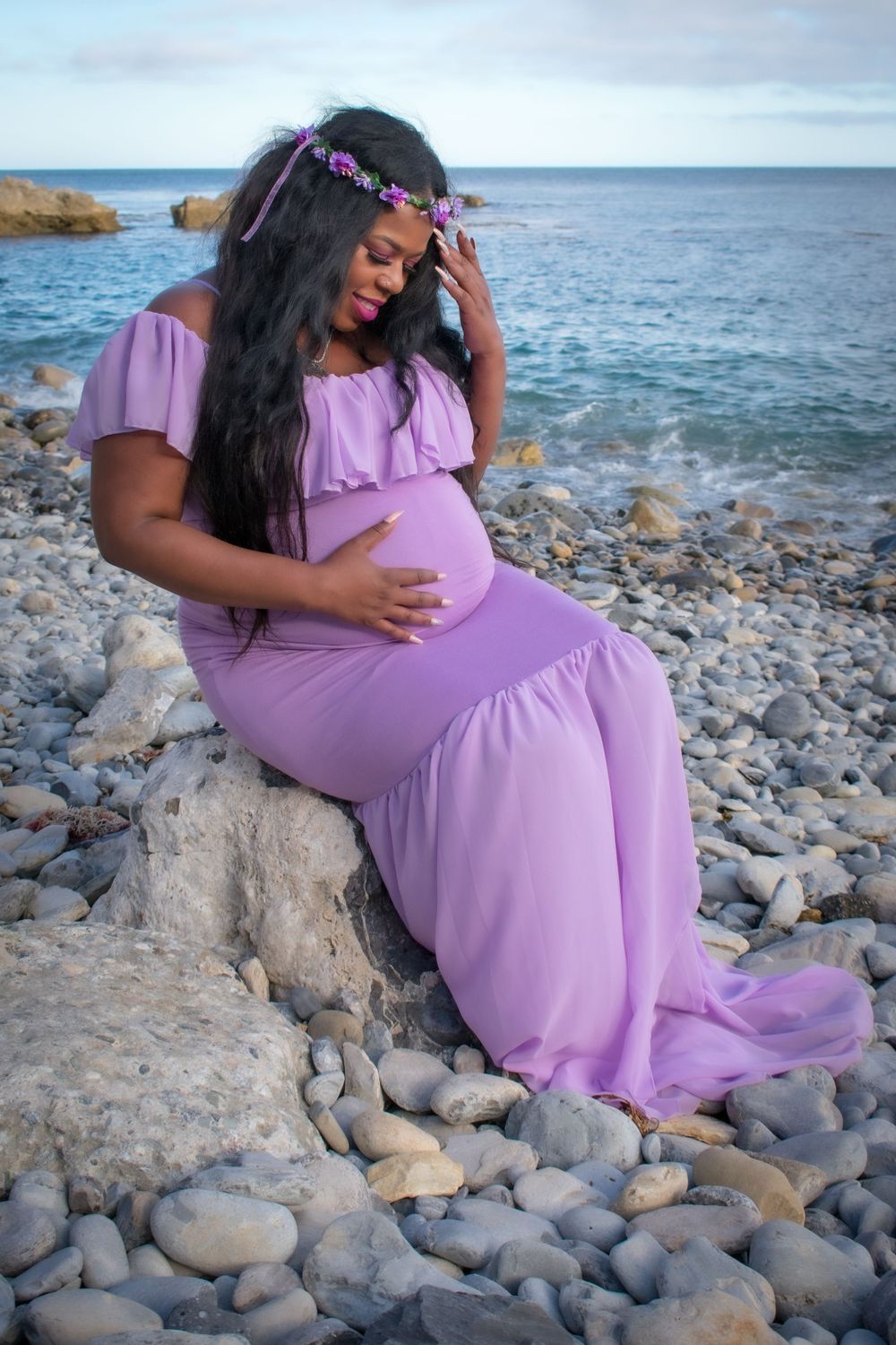 Pregnant woman in purple maternity gown sits on rocky beach holding baby bump near ocean.
