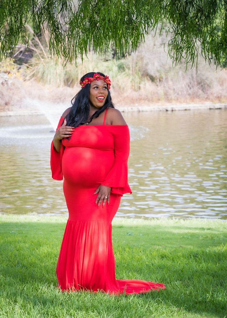 Pregnant woman in red off-shoulder gown with floral crown poses by a pond in a maternity photoshoot.