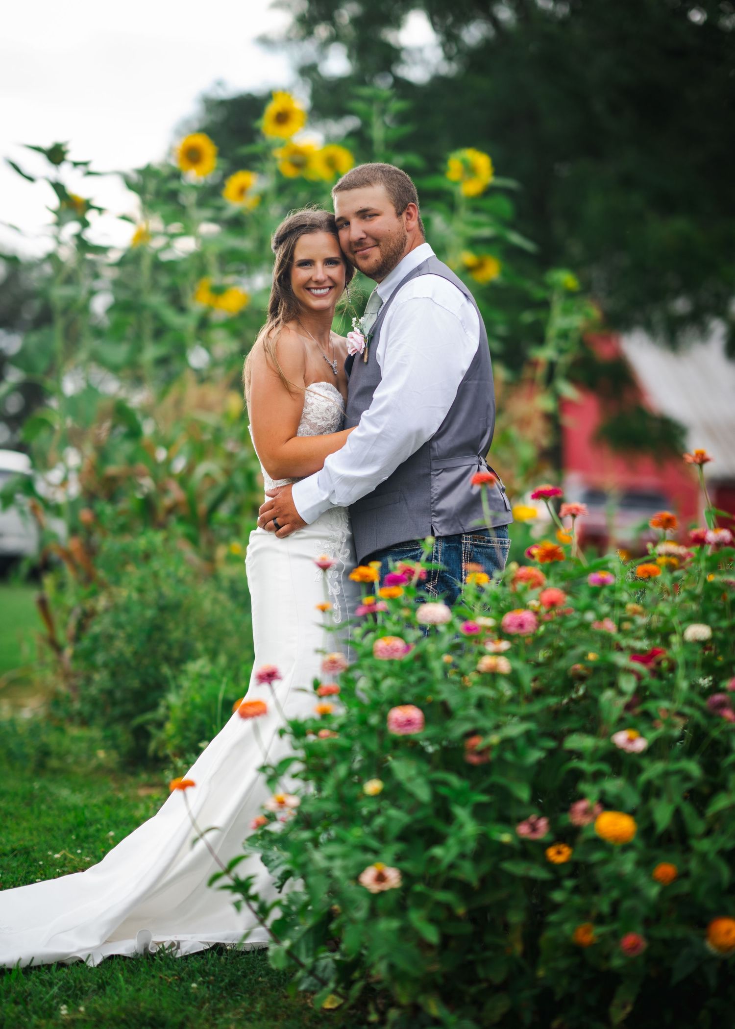 A couple poses in a garden with bright sunflowers and colorful flowers in the background.