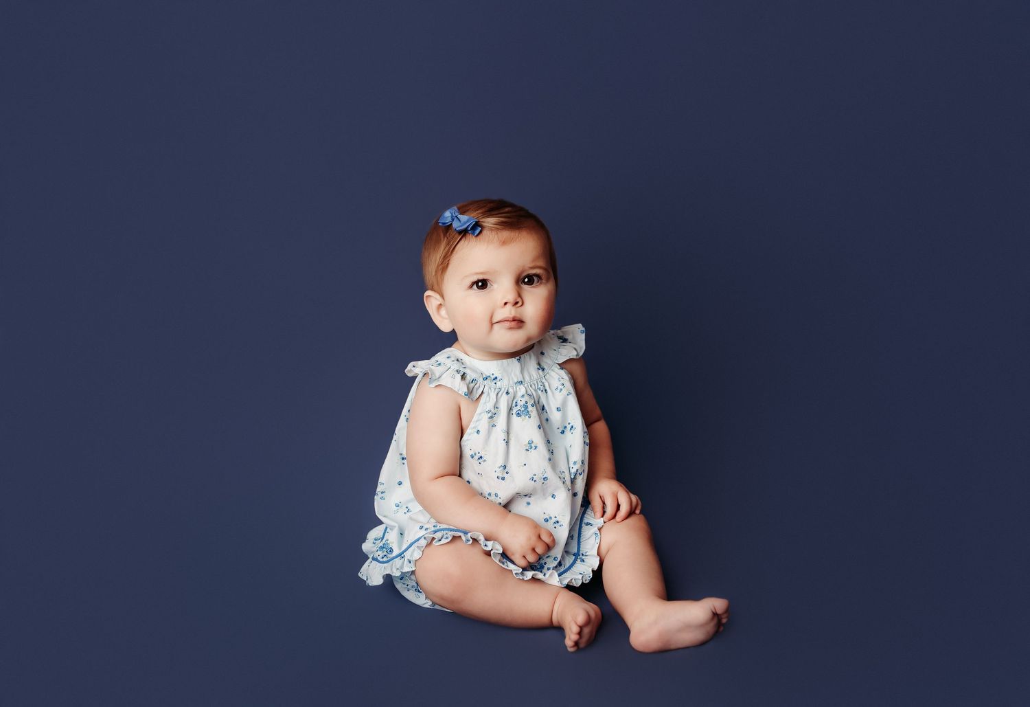 baby girl getting her 9 moth pictures taken in tulsa ok. Baby girl is in a blue floral dress on a solid blue background