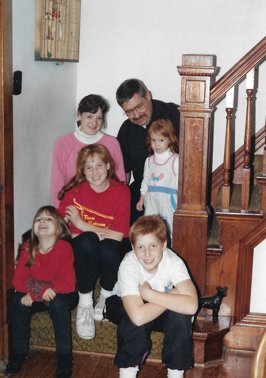 A family portrait on wooden stairs inside a home with traditional banister and wall decor.