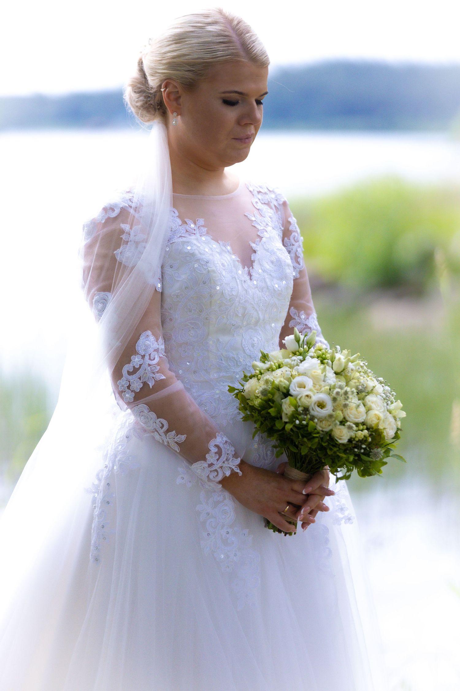 Blonde bride in elegant lace long-sleeve wedding dress holding bouquet of white flowers by a lake.