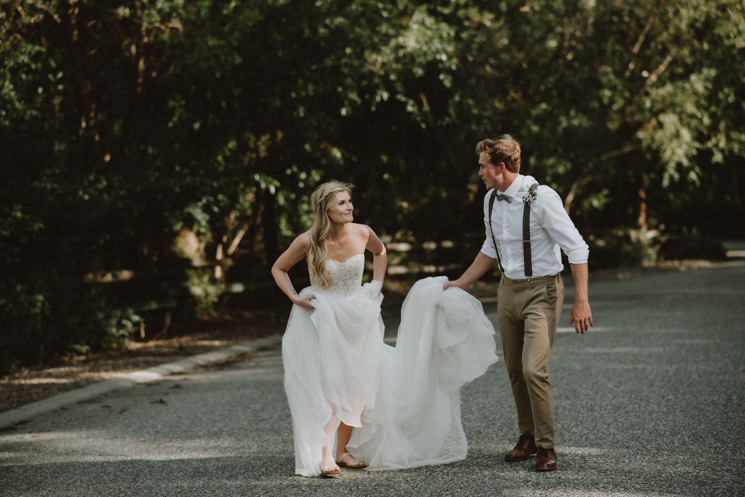 Playful couple walking on road while bride holds up wedding dress.