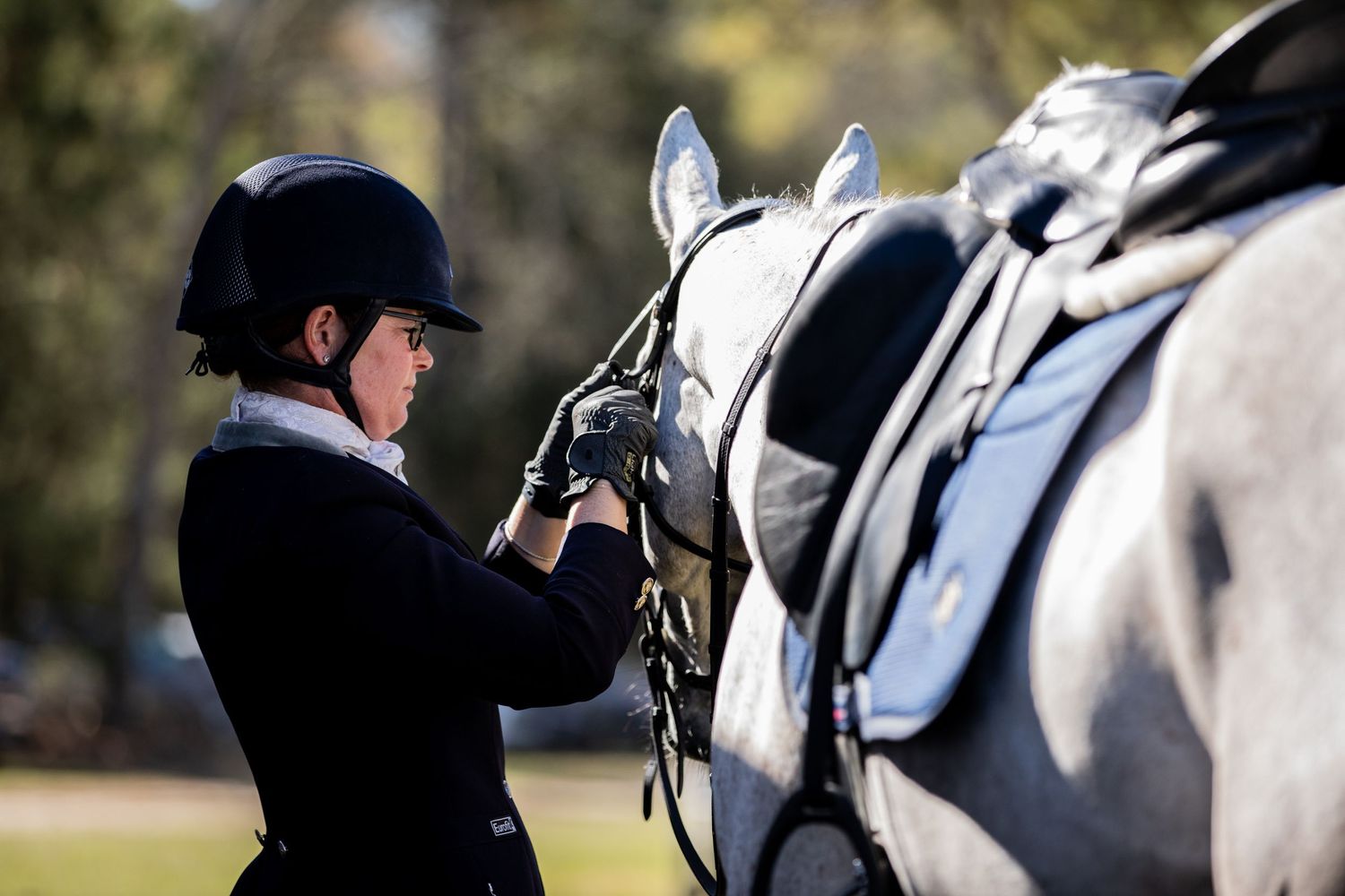 Equestrian adjusting tack on gray horse before competition in outdoor setting.