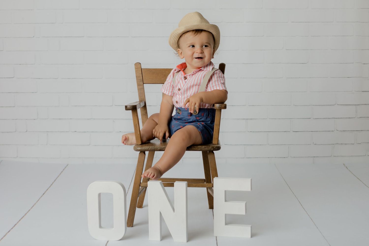 One year studio portrait for a little boy sitting in a wooden chair and a brick background while smiling.