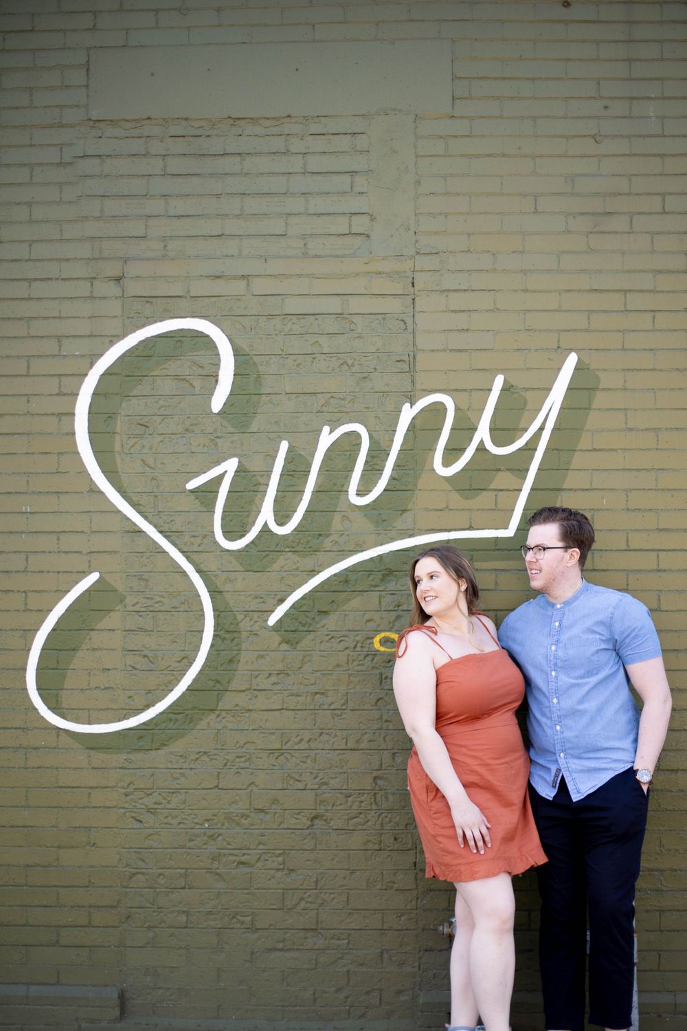 A couple poses against a brick wall with stylized 'Sunny' text artwork.