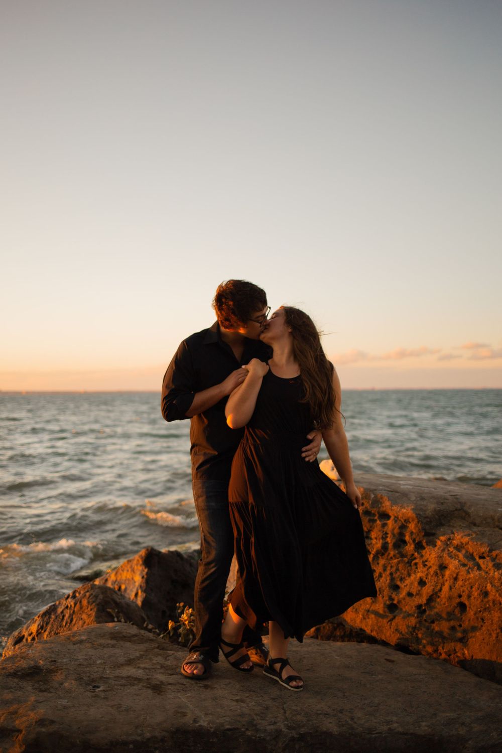 A couple shares a romantic moment at sunset on a rocky oceanside cliff.
