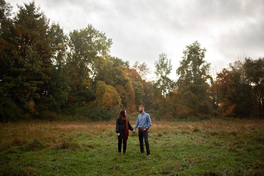 A couple walks hand in hand through an autumn field with trees in the background.
