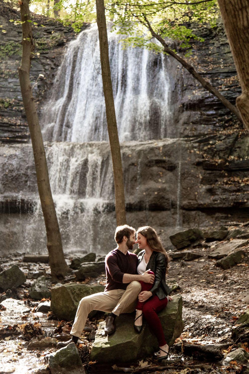 A couple sits on rocks in front of a majestic waterfall in a forest setting.