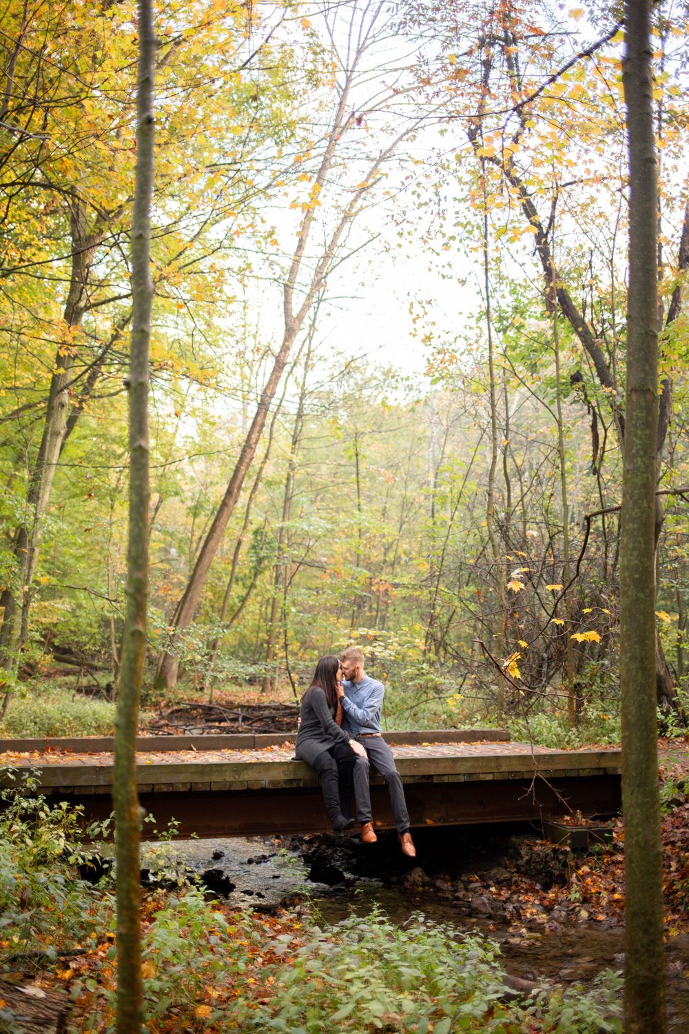 A couple sits together on a wooden bridge surrounded by autumn foliage.