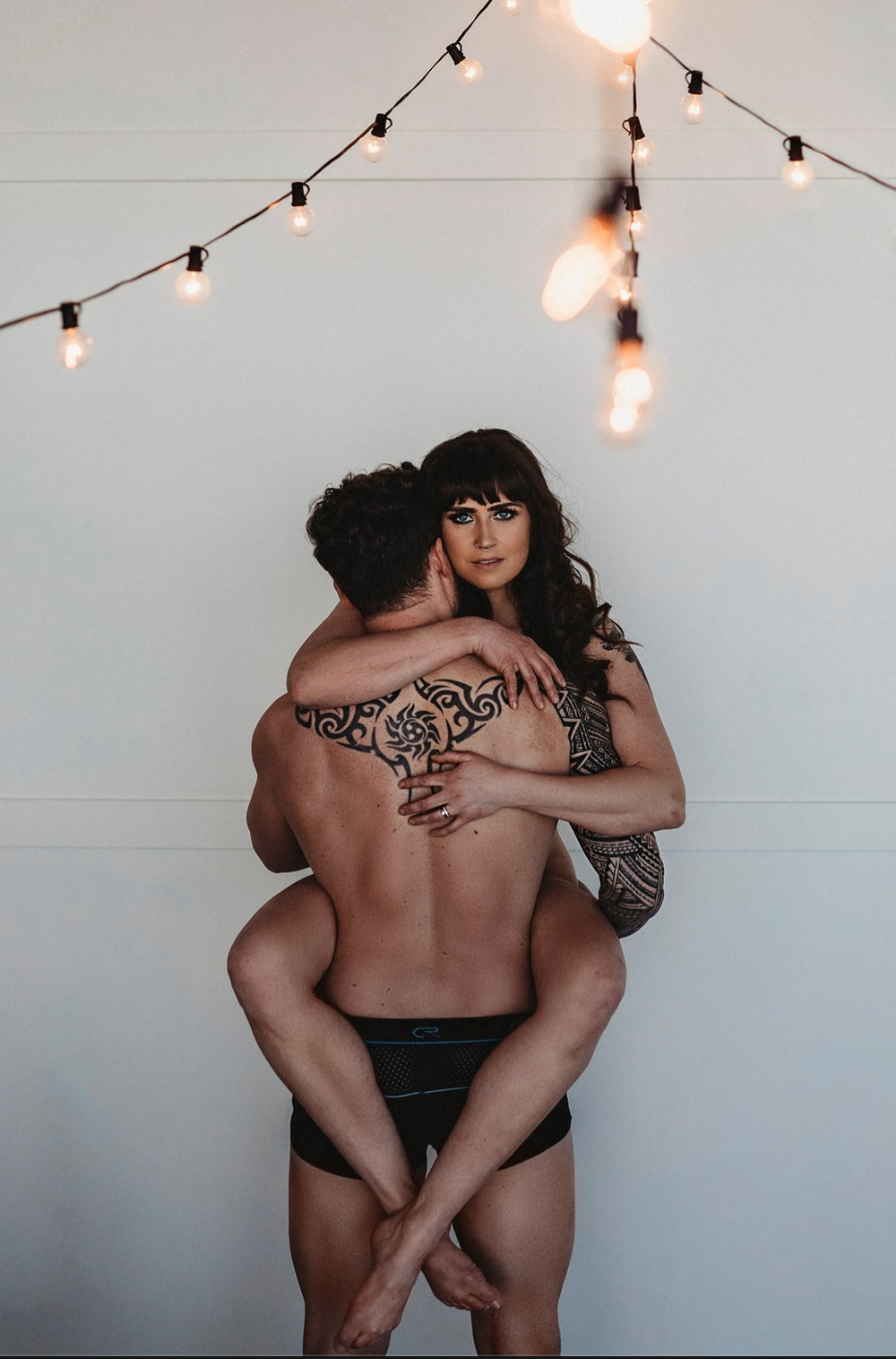 Intimate couple embrace under string lights in artistic boudoir photo.