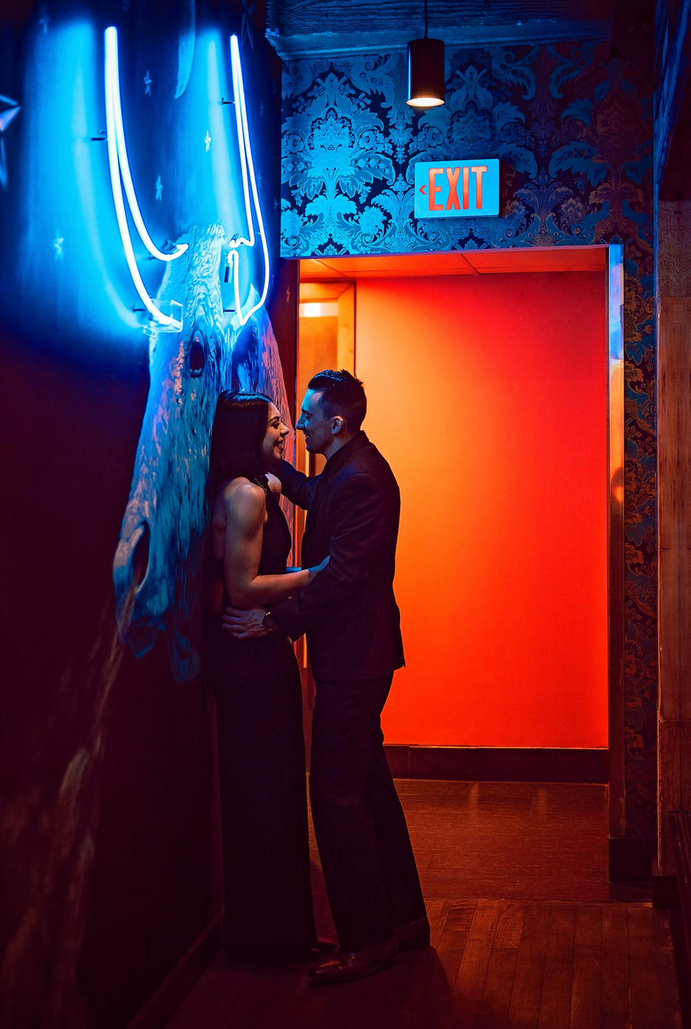 Couple in formal wear embracing in front of neon lights and orange doorway.
