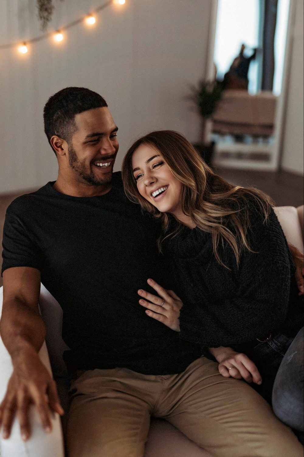 Couple laughing together on couch under warm string lights.