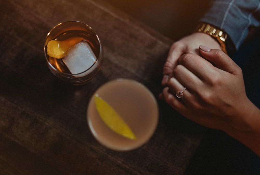 Two cocktails on a dark wooden surface with a ring-adorned hand visible.