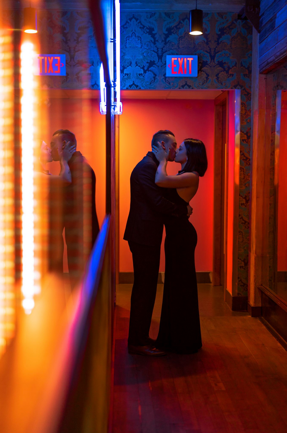 A romantic silhouette of a couple in formal wear against neon lights near an exit sign.