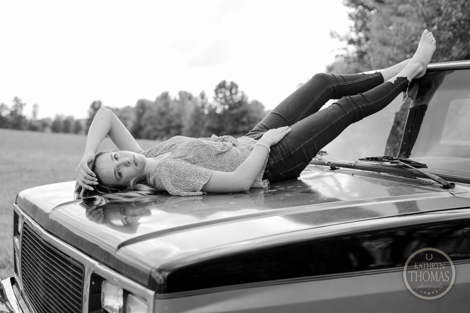 Black and white photo of someone reclining on the hood of a vintage truck.