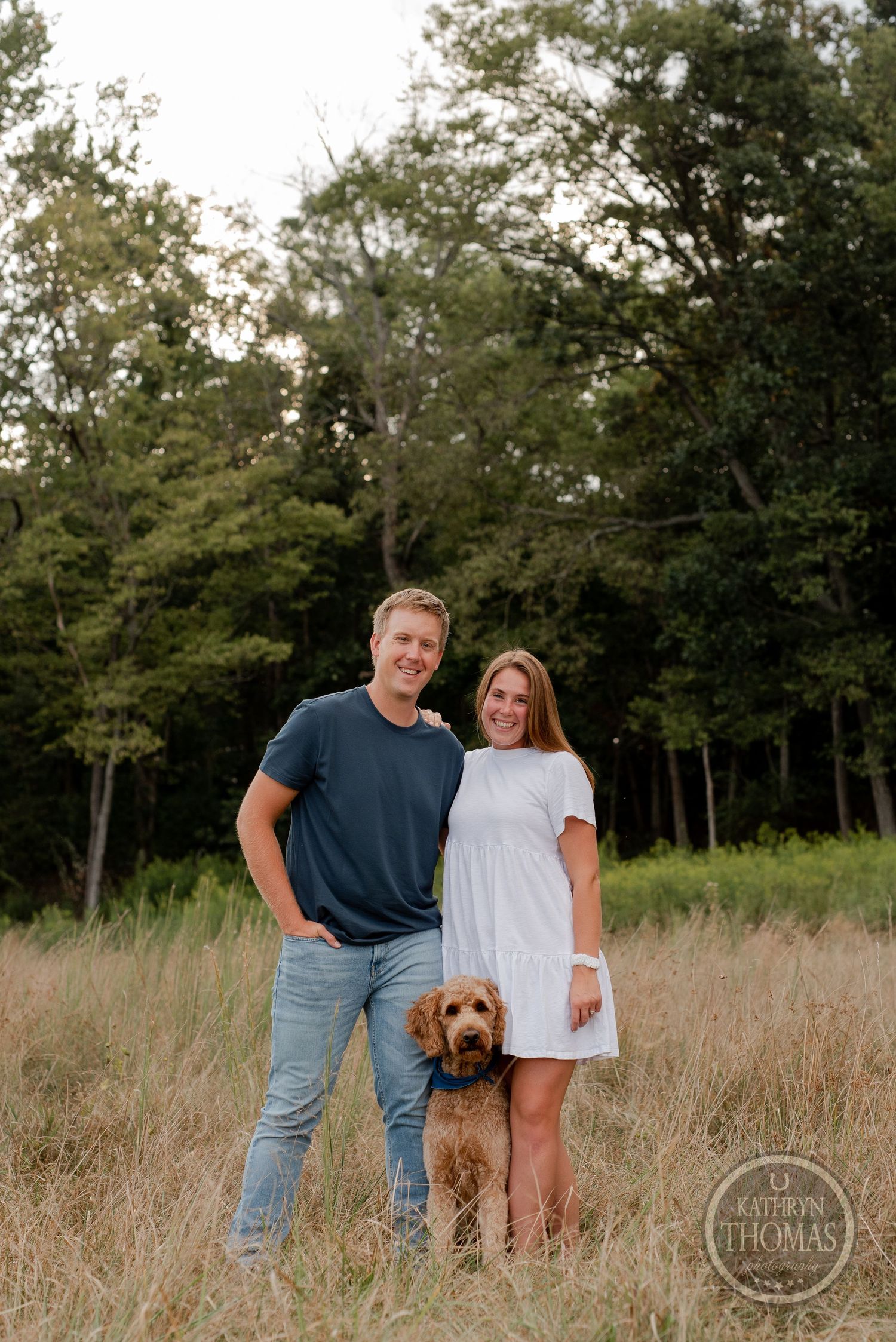 A couple and their goldendoodle dog pose for a portrait in a rustic field during golden hour.