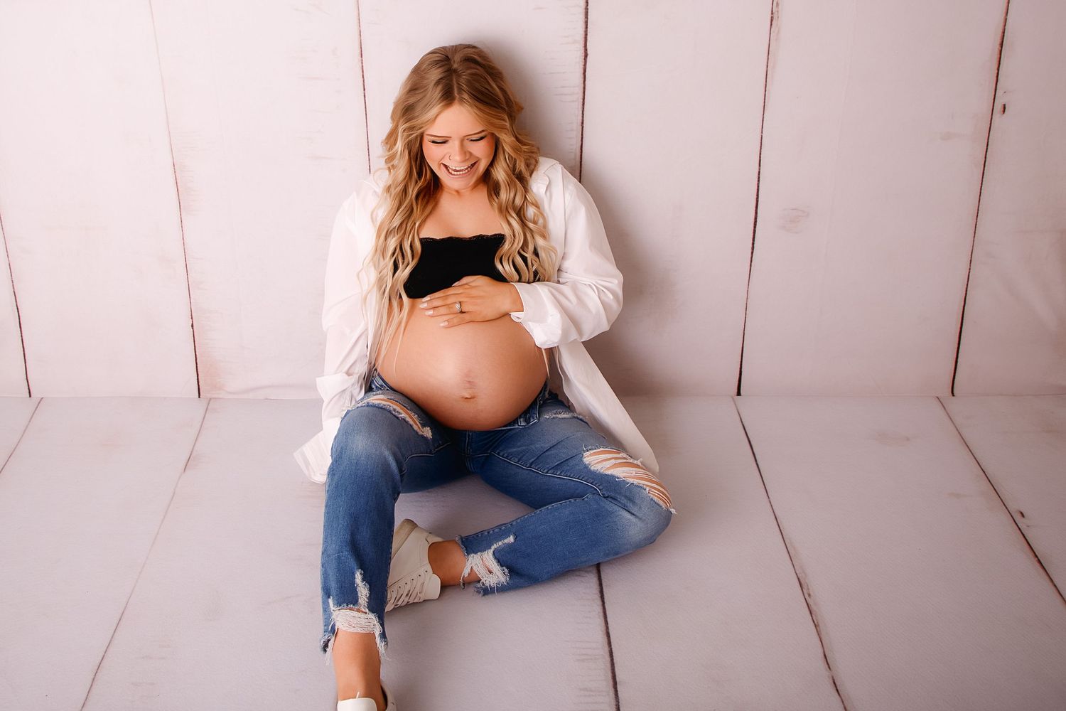 Expectant mother in jeans and cardigan sits on white wooden floor during maternity photoshoot.