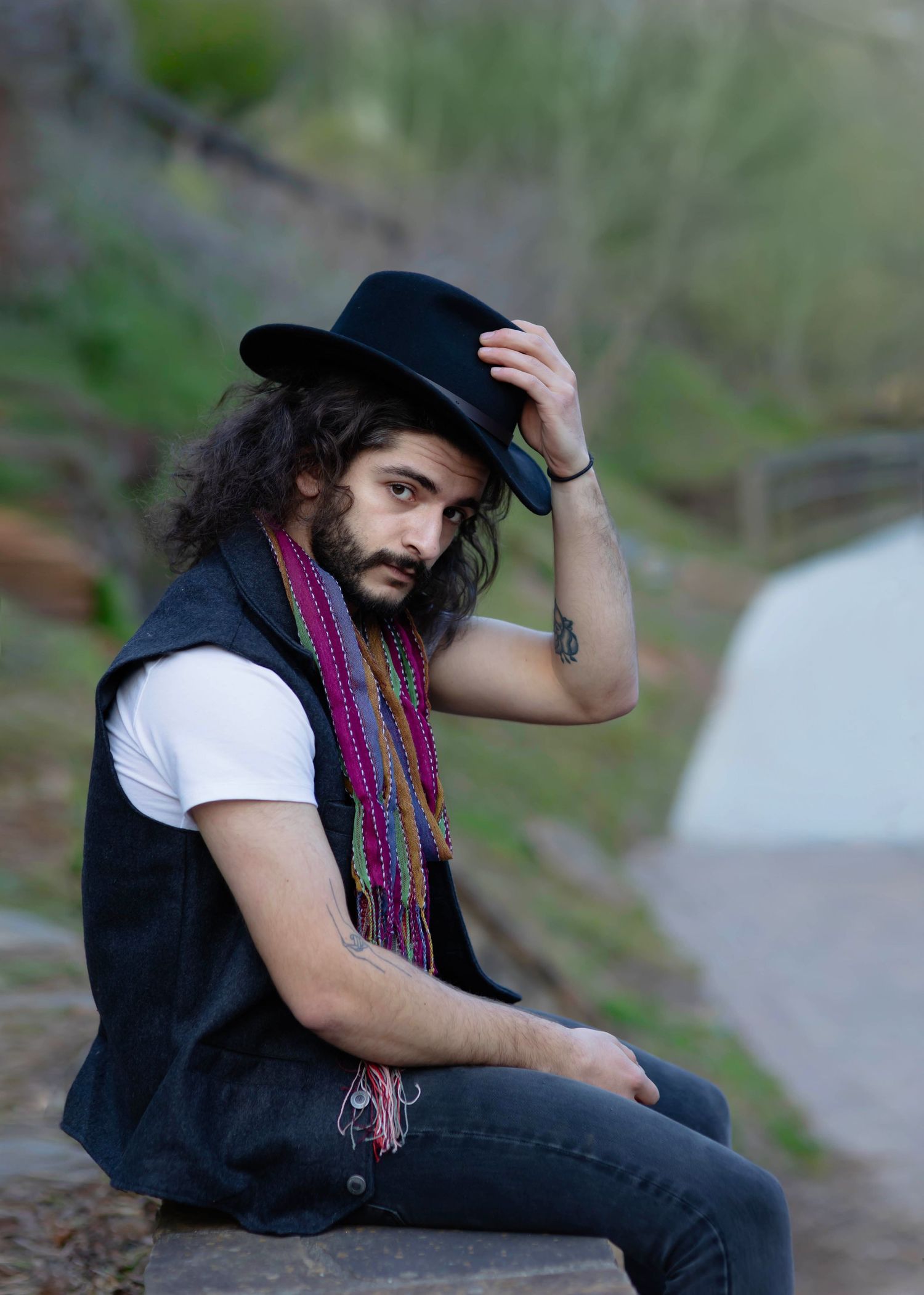 Young man with tattoos holding his hat with his left hand on his head sitting on the stones