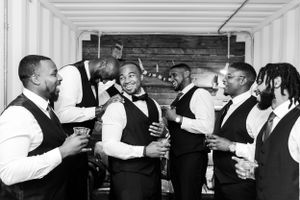 A group in formal attire wearing black vests and bowties laugh and socialize while holding drinks at an event.