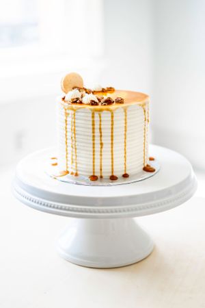 Elegant white cake with caramel drip and cookie decorations on a ceramic cake stand.
