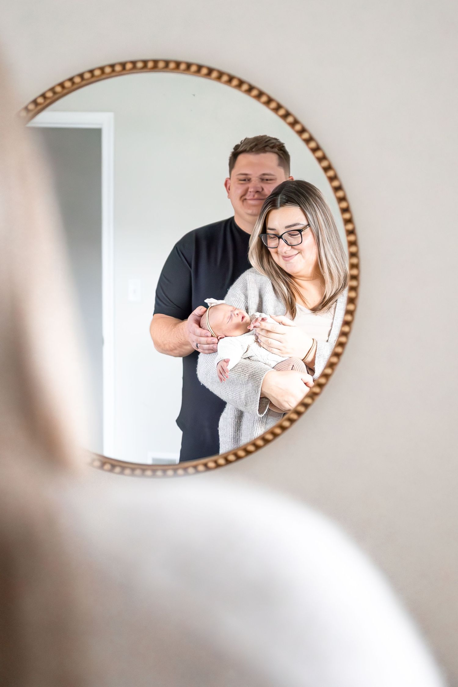 A couple shares a tender moment with their newborn baby in front of a round gold mirror.