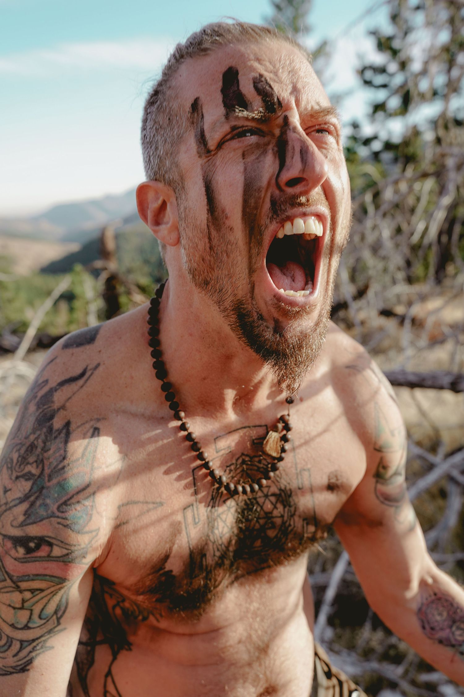 Shirtless tattooed person showing intense emotional expression outdoors.