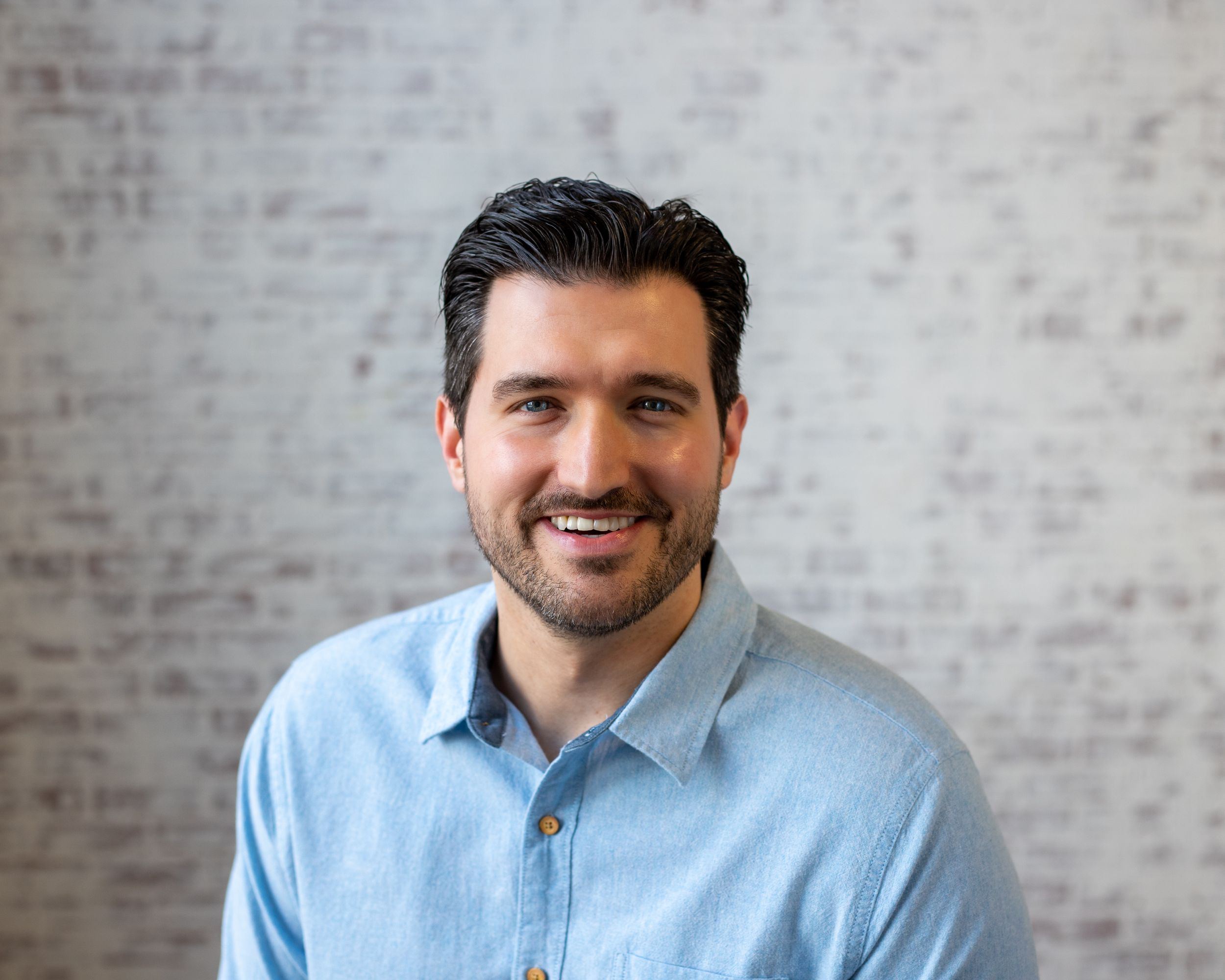 Professional headshot of person in light blue button-down shirt smiling against a grey textured wall background.