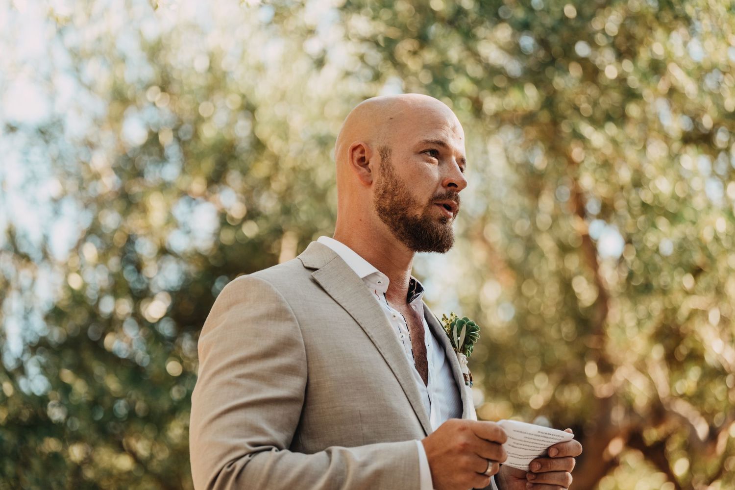 A person in a beige suit holds papers while speaking during an outdoor ceremony.