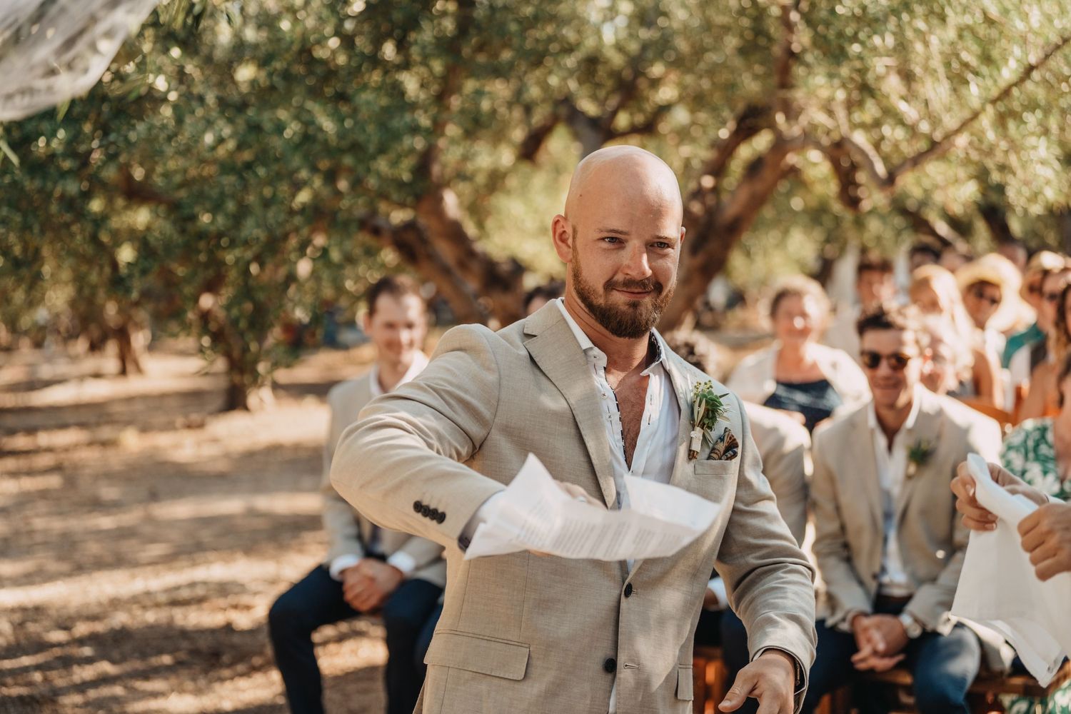 A person in a light suit gestures expressively during an outdoor wedding ceremony.