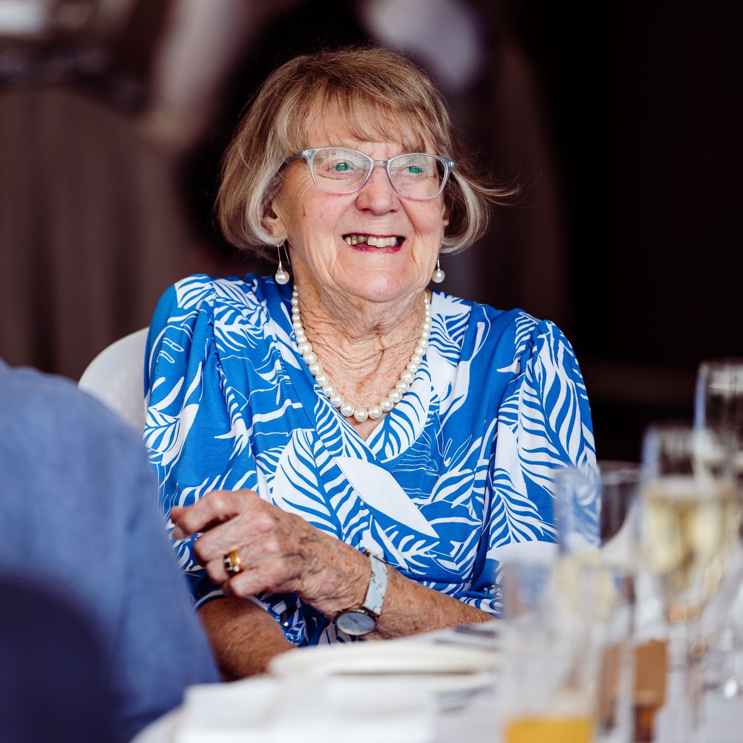 Guest wearing a blue floral top and glasses laughs during a wedding reception.