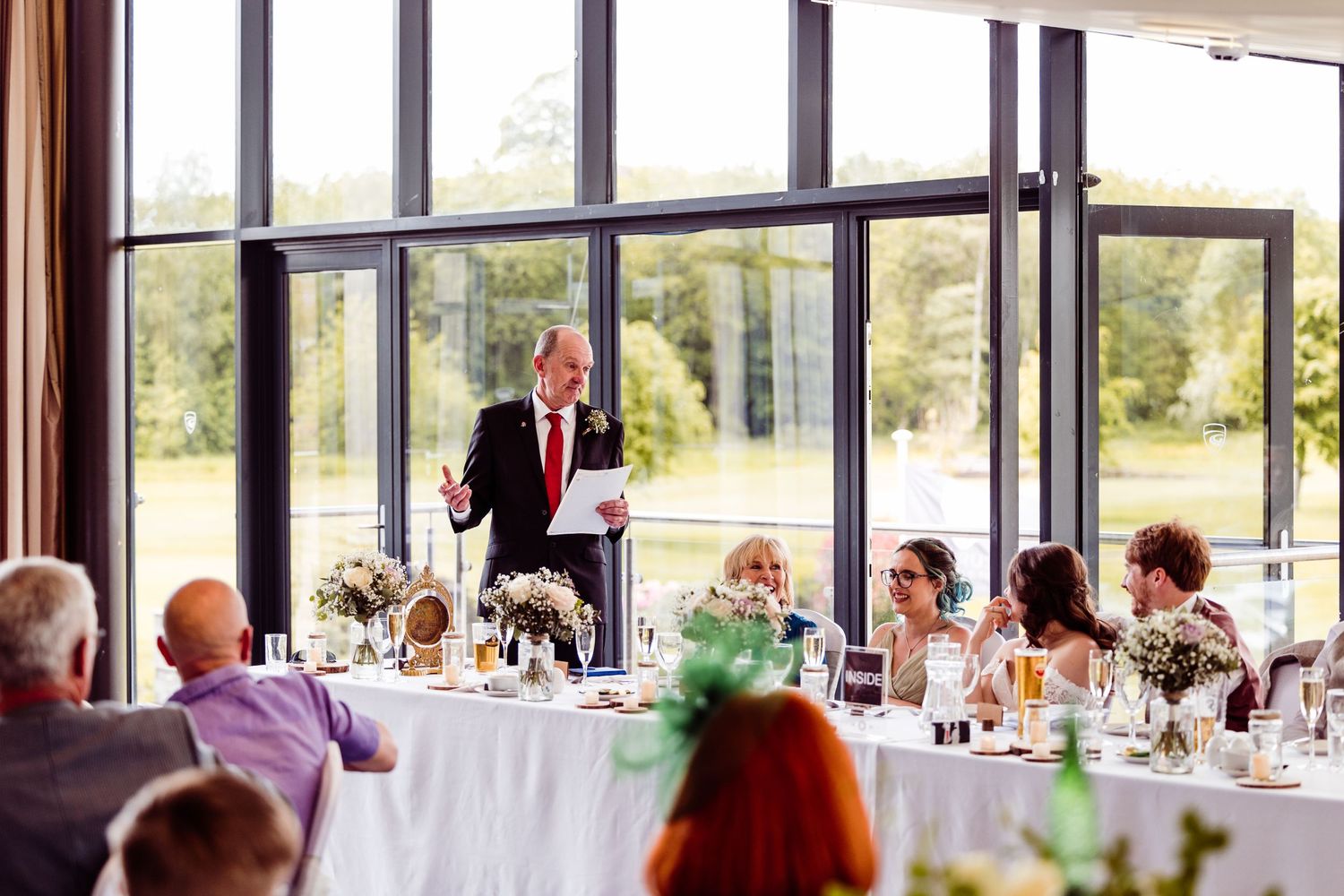 A speaker in red tie addresses guests during a wedding reception speech in a bright venue.