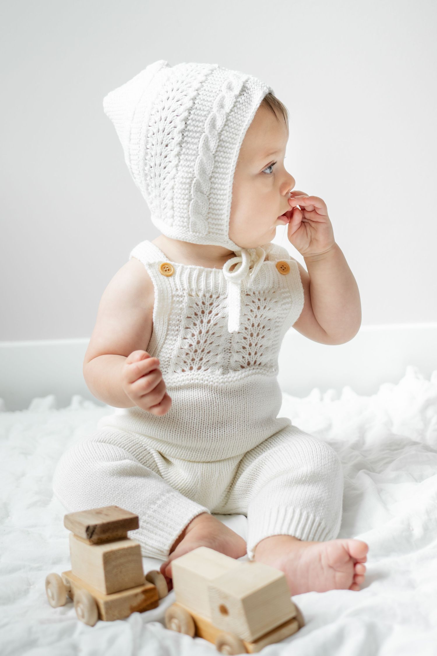 little boy in white knit two-piece outfit puts hands in his mouth and looks over his left shoulder