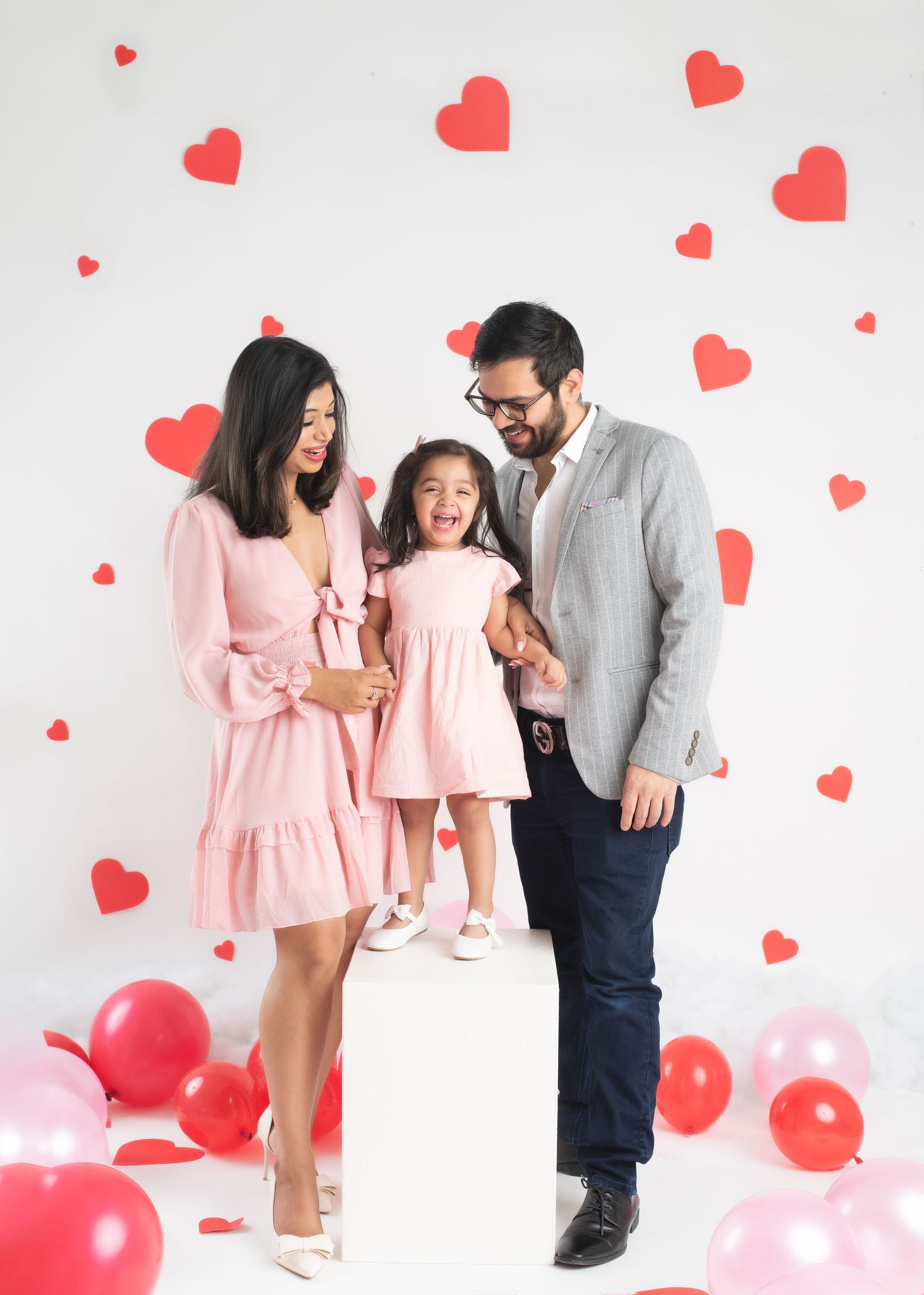 a family of three stand in front of a white backdrop with red hearts and smile for their minneapolis family photographer